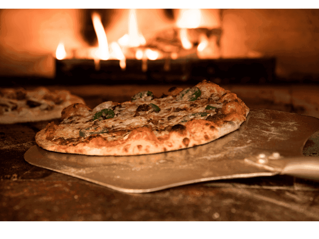 Pizza artisanale en cours de cuisson dans un four à bois, posée sur une pelle enfarinée, avec croûte dorée et garniture généreuse, illustrant la rotation manuelle pour une cuisson homogène