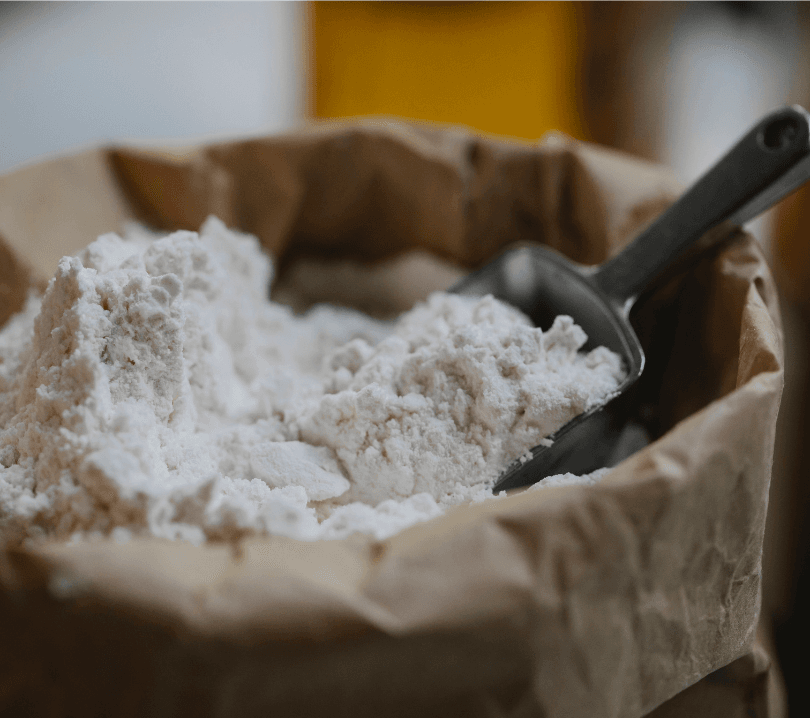 Gros plan sur un sac en papier kraft rempli de farine pour pate pizza, avec une cuillère en métal enfoncée dans la farine.
