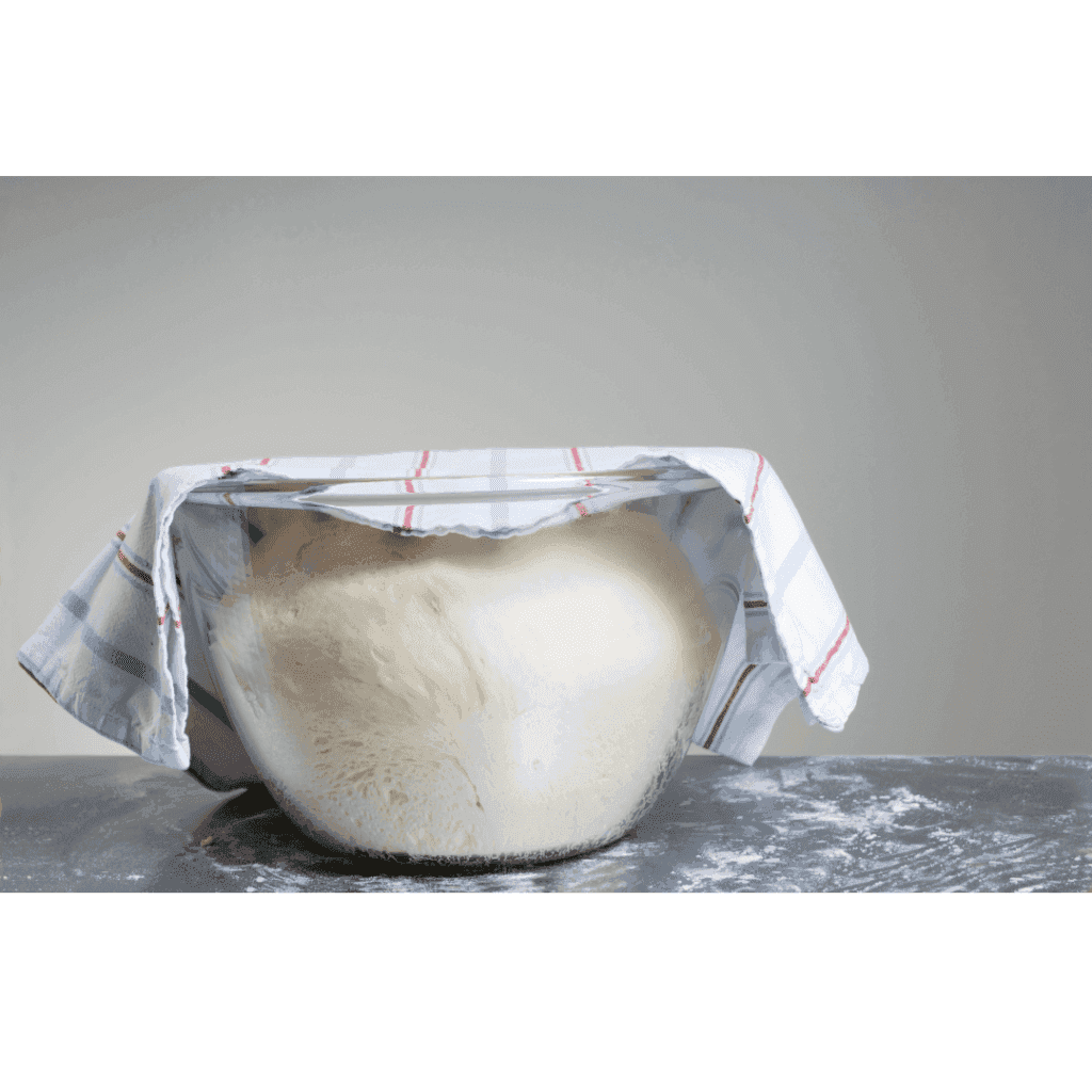 Pâte à pizza qui lève dans un saladier en verre recouvert d’un torchon, sur un plan de travail fariné