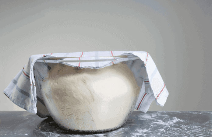 Pâte à pizza qui lève dans un saladier en verre recouvert d’un torchon, sur un plan de travail fariné