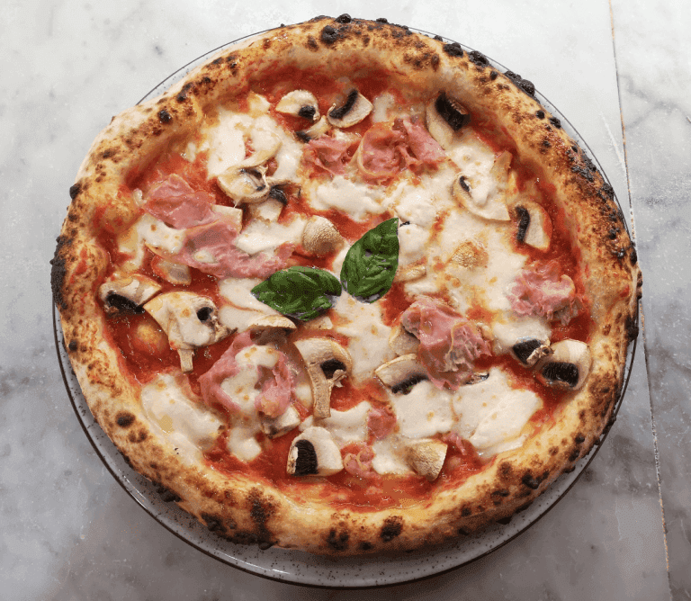 Pizza royal garnie de jambon, champignons frais, mozzarella fondue et feuilles de basilic, servie sur une assiette en marbre