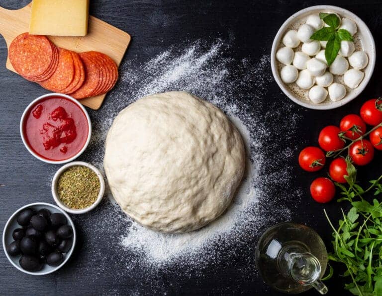 Boule de pâte biga entourée d’ingrédients frais pour pizza : tomates cerises, mozzarella, sauce tomate, origan et olives noires