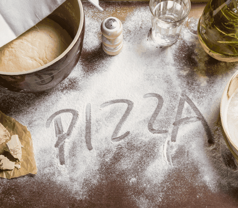 Mot “PIZZA” tracé à la main dans de la farine sur un plan de travail, entouré de levure fraîche, d’huile d’olive, d’un saladier de pâte levée et de farine tamisée.