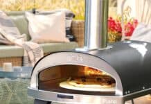 Four à pizza ZiiPa Piana en fonctionnement sur une table de terrasse, pizza en cuisson à l’intérieur avec flammes visibles et salon de jardin flou en arrière-plan.