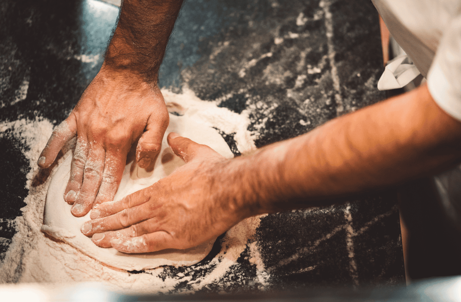 Deux mains farinées tiennent de la farine au-dessus d’un plan de travail saupoudré, en pleine préparation culinaire.