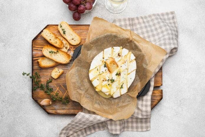 Camembert rôti au four entaillé et fondu, servi avec tranches de pain grillé, raisins et herbes, sur plateau en bois