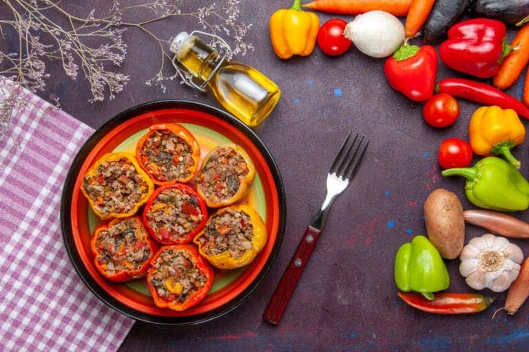 Vue de dessus d’un plat coloré de poivrons farcis au four, entouré d’ingrédients crus comme carottes, ail, oignons, tomates et poivrons pour illustrer la farce pour poivron.