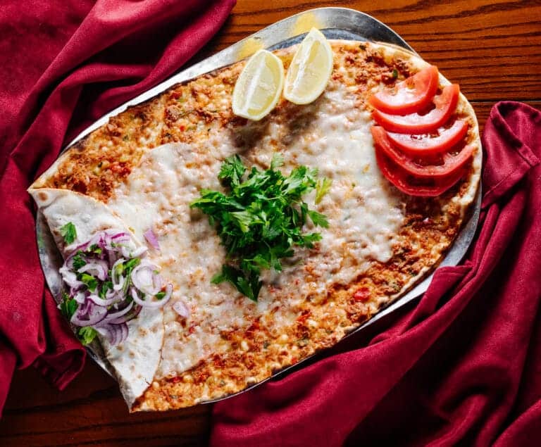 Lahmacun garni de fromage fondu, tomates, oignons et persil, entouré d’un tissu rouge sur fond de bois