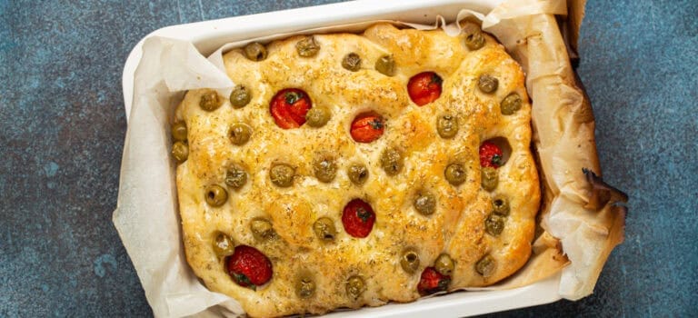 Focaccia italienne cuite au four avec olives vertes, tomates cerises et huile d’olive, à la surface dorée et pleine de creux caractéristiques.
