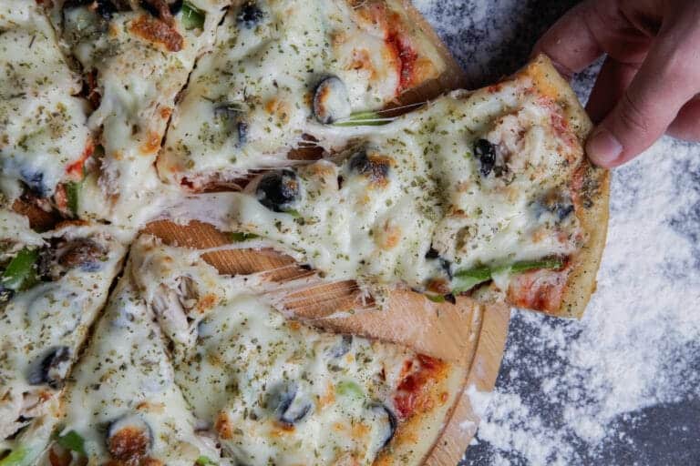 Une main saisit une part de pizza romaine croustillante, garnie de fromage fondu, olives noires et légumes, sur fond de farine.