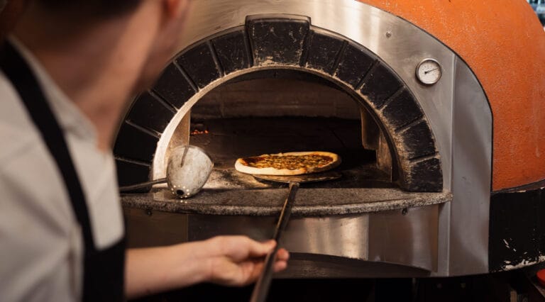 Pizza insérée dans un four pizza professionnel à bois montrant la sole chauffée et la flamme active.