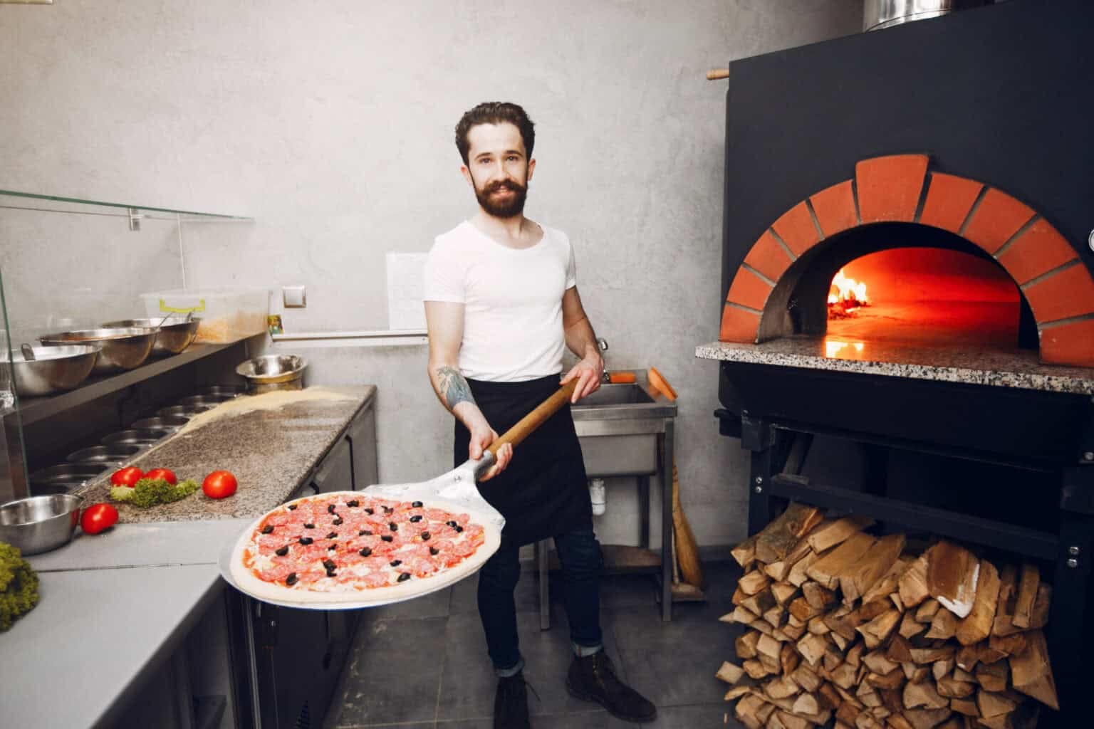 Cuisinier tenant une pizza sur une pelle face à un four professionnel à bois en pleine chauffe.