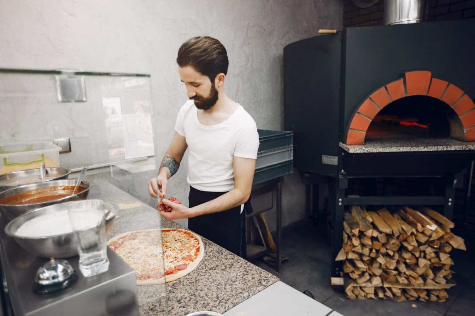 Pizzaïolo garnissant une pizza devant un four pizza professionnel à bois chauffé pour la cuisson.