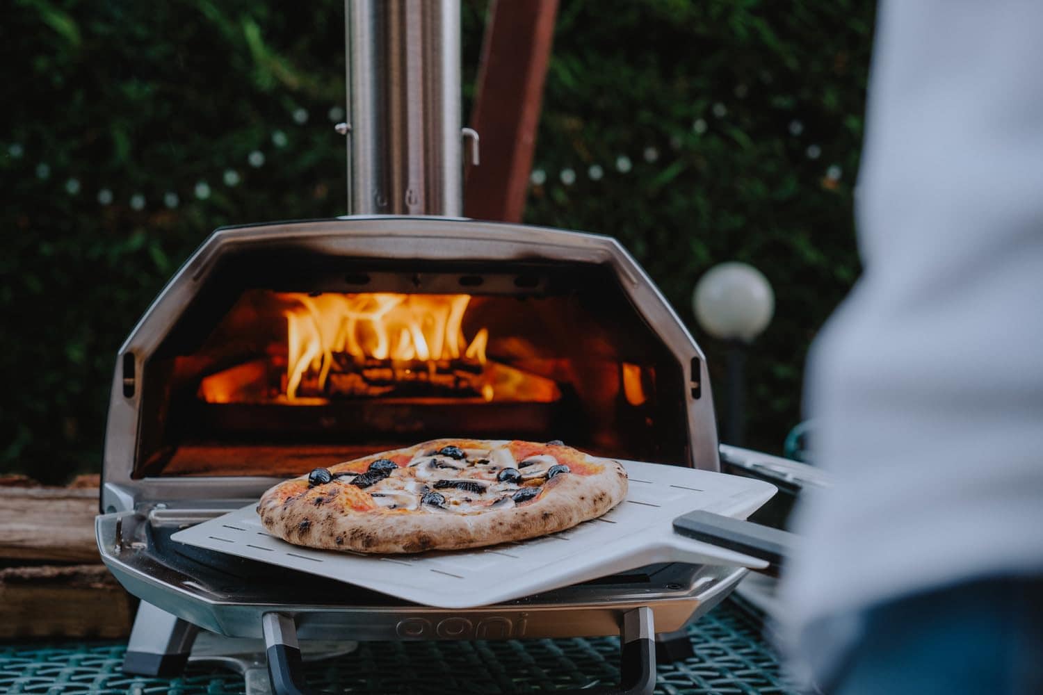 Pizza en cours de cuisson dans le Ooni Karu 16 avec flamme à l’intérieur et pelle à pizza au premier plan