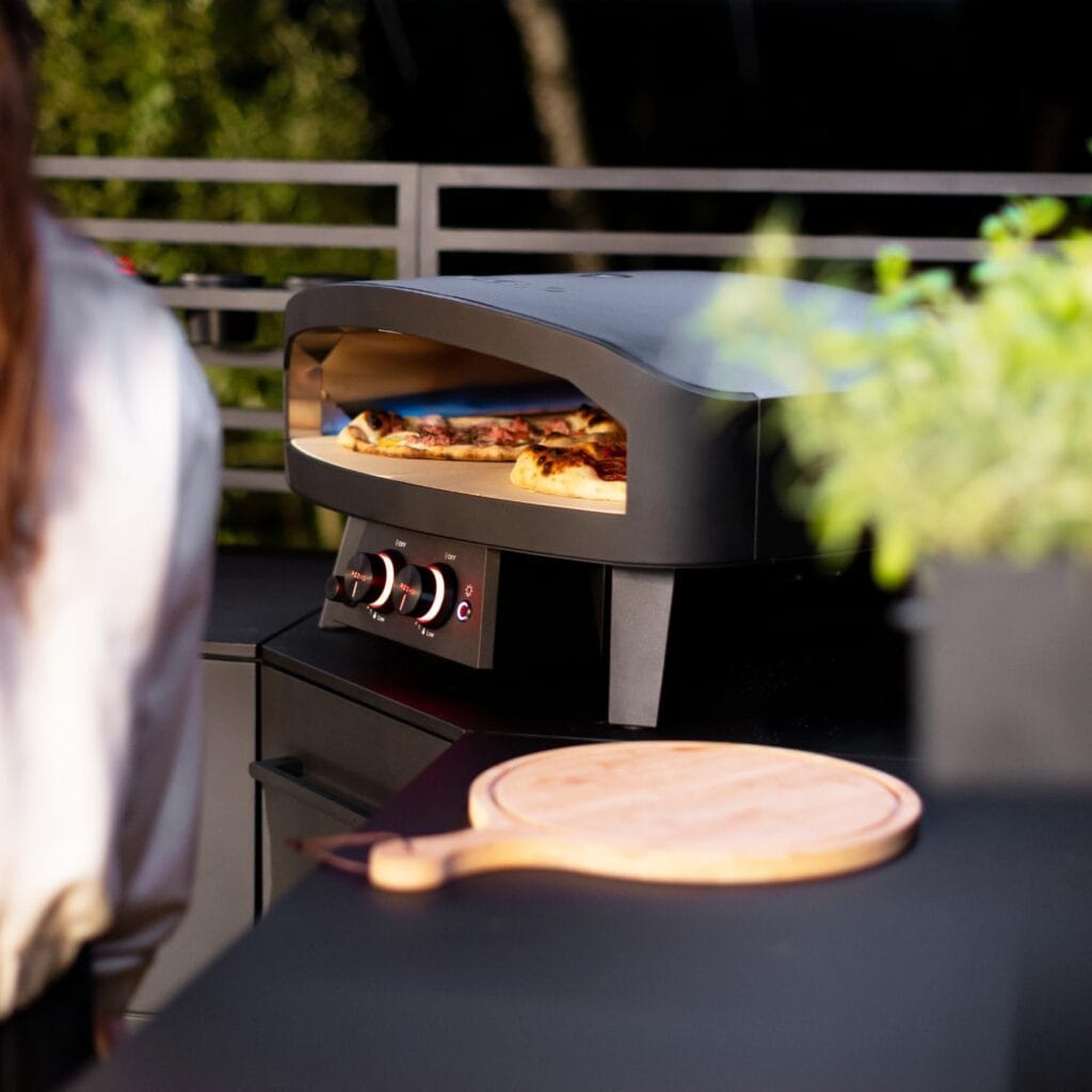 Four pizza Cozze 24" Black Edition : l'excellence du gaz installé sur une terrasse avec pizza en cuisson dans un four extérieur moderne.