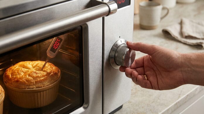 Main ajustant le bouton d'un four en inox avec un ramequin de pâtisserie et un thermomètre à l'intérieur.