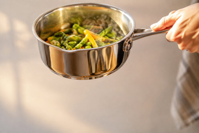 Casserole Fissler en inox avec légumes sautés Une main tient une casserole Fissler en acier inoxydable brillant remplie de légumes verts et jaunes cuits.