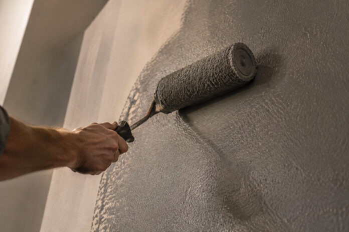 Une main utilisant un rouleau à peinture pour appliquer une couche de peinture grise sur un mur.
